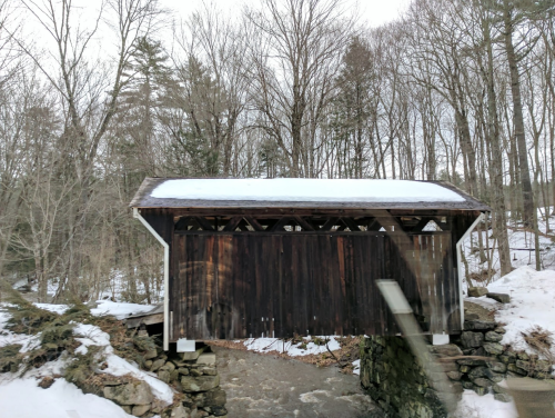 A wooden covered bridge with a snow-covered roof, surrounded by trees and a snowy landscape.