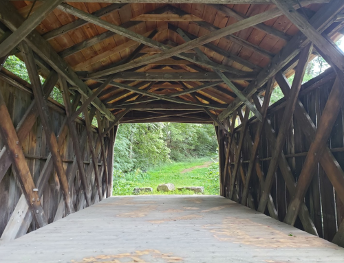 View from inside a wooden covered bridge, showcasing a rustic interior and greenery beyond the entrance.