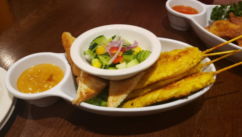 A plate featuring chicken satay skewers, a fresh salad, toasted bread, and a side of peanut sauce.