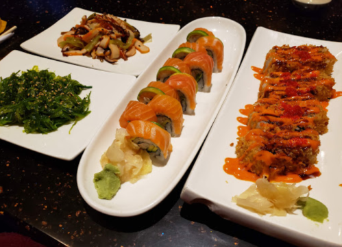 A variety of sushi dishes on white plates, including rolls, tempura, and seaweed salad, garnished with wasabi and ginger.