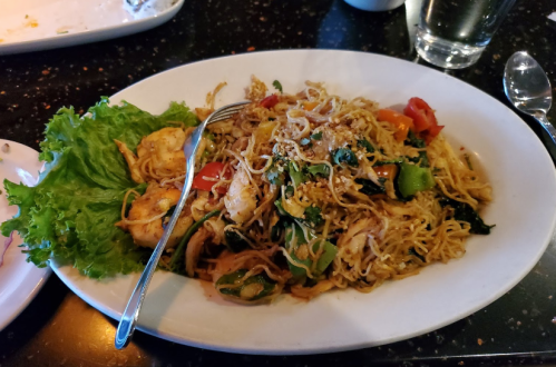 A plate of stir-fried noodles with shrimp, vegetables, and a side of lettuce on a dark table.