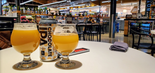 Two glasses of beer sit on a table with a can of Chocolate IPA in a vibrant grocery store setting.
