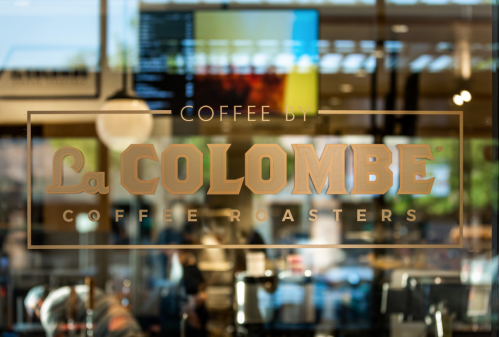 Sign for La Colombe Coffee Roasters displayed on a glass window, reflecting a busy café interior.