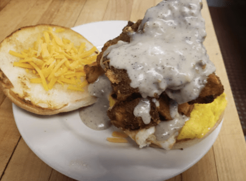 A plate featuring a biscuit sandwich with fried chicken, gravy, and shredded cheese on the side.