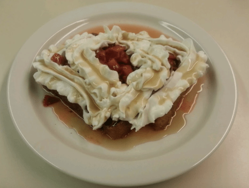 A plate of dessert featuring a slice of cake topped with whipped cream, strawberries, and a drizzle of syrup.