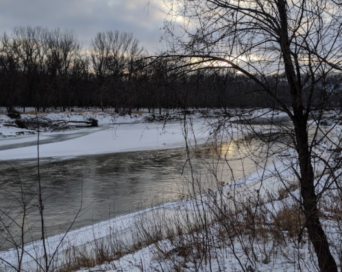 A serene winter scene of a river with ice, surrounded by bare trees and a cloudy sky.