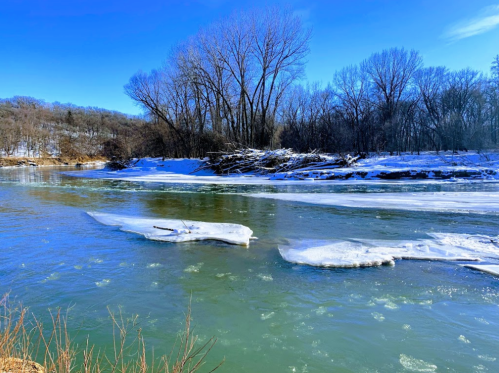 A serene river scene with ice patches, snowy banks, and bare trees under a clear blue sky.