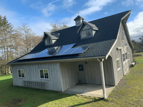 A modern barn-style house with solar panels on the roof, surrounded by greenery and trees under a clear blue sky.