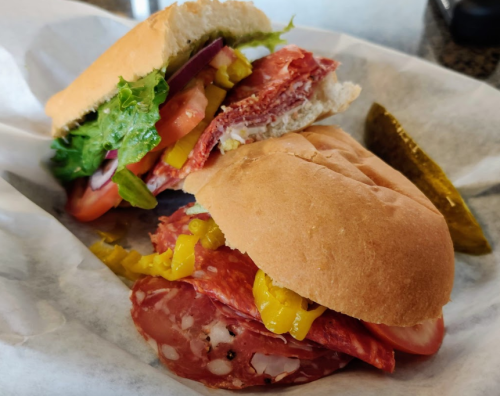 A close-up of two sandwiches with salami, lettuce, tomato, and pickles on a white paper background.