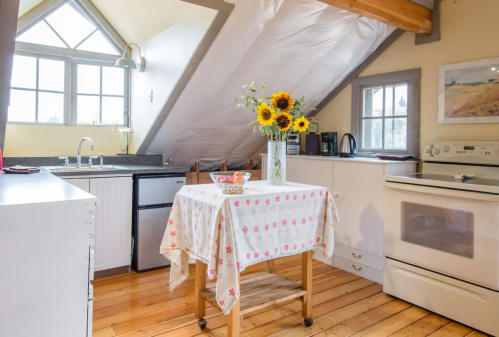 Cozy kitchen with wooden floors, a table with a floral tablecloth, sunflowers, and a sloped ceiling.