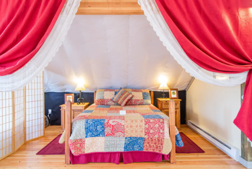 Cozy bedroom with a quilted bedspread, draped red curtains, and warm lighting in a tent-like setting.