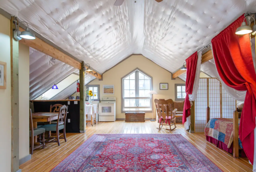 Cozy attic space with wooden beams, a red rug, dining area, kitchenette, and a bed, featuring soft lighting and decor.