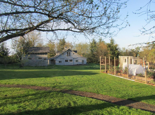 A sunny farm scene featuring a rustic barn, garden beds, and lush green grass surrounded by trees.