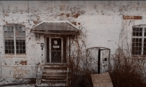 Abandoned building with peeling white paint, overgrown vines, and a weathered door, creating a haunting atmosphere.