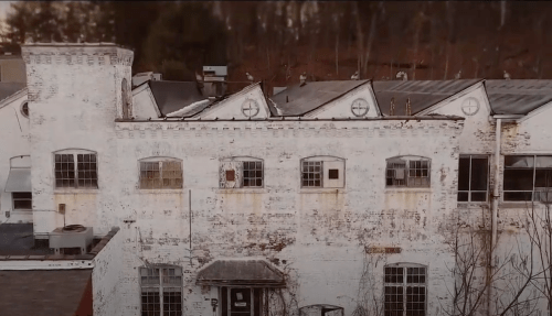 Aerial view of an old, weathered brick building with multiple windows and a sloped roof, surrounded by trees.