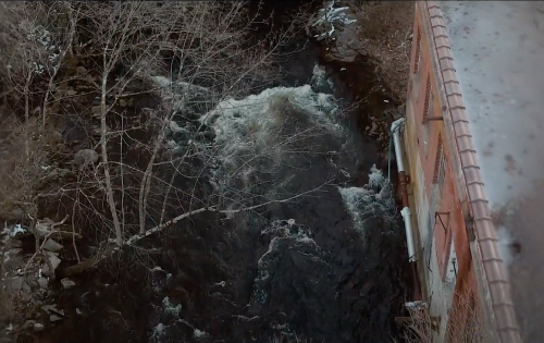 Aerial view of a flowing river beside a building, with bare trees and rocky banks visible.