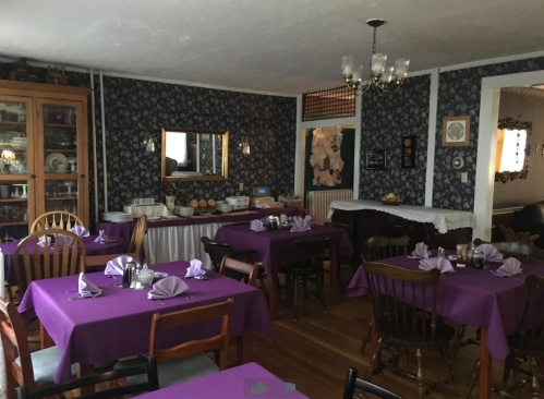 Cozy dining area with purple tablecloths, wooden chairs, and floral wallpaper, set for a meal.
