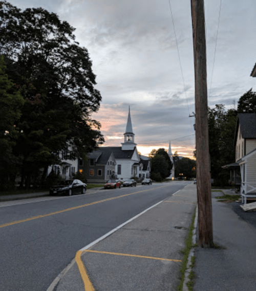 A quiet street lined with trees and houses, featuring a church steeple against a sunset sky.