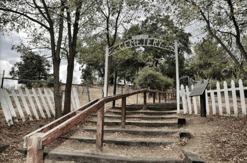 A weathered wooden staircase leads to a cemetery entrance, framed by trees and a white picket fence.