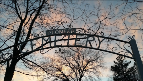 Sign reading "Pioneer Cemetery" against a colorful sunset sky, framed by bare trees.