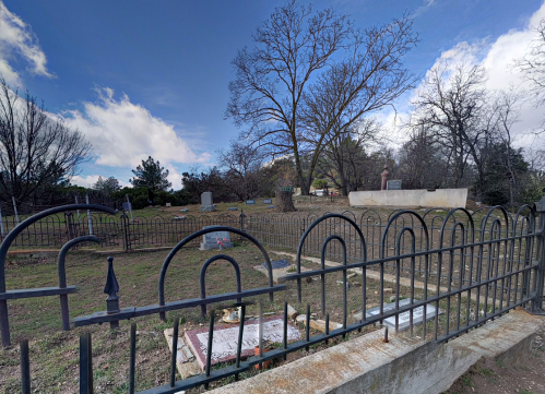 A cemetery scene with a wrought iron fence, gravestones, and a large tree under a partly cloudy sky.