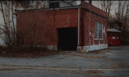 Abandoned red brick building with a dark entrance, surrounded by overgrown weeds and cracked pavement.