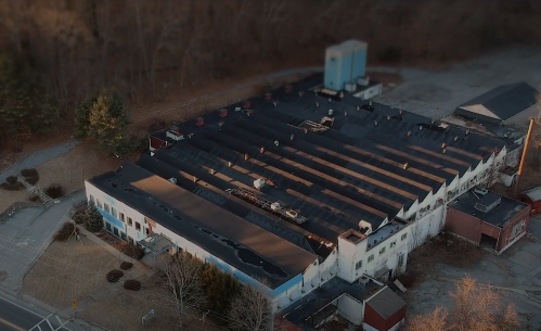Aerial view of a large, industrial building with a flat roof, surrounded by trees and empty land.