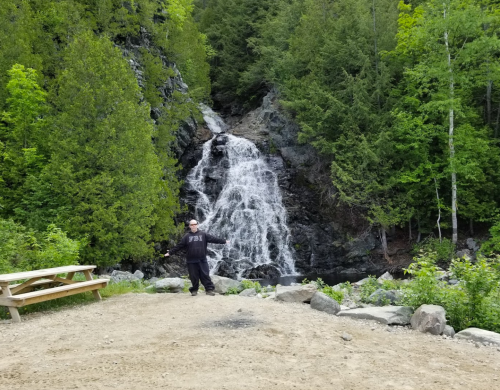 A person in an FBI jacket stands near a waterfall surrounded by lush green trees and rocky terrain.