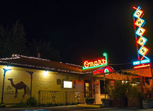 A brightly lit restaurant sign reading "Omar's" at night, with decorative lights and a camel mural on the building.
