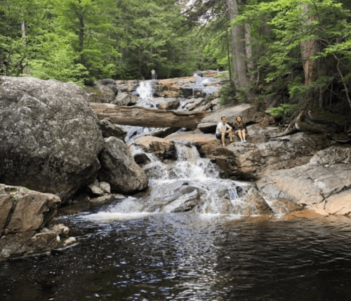 A serene waterfall cascades over rocks, with lush greenery surrounding a couple sitting by the water's edge.