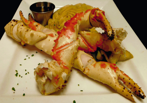 A plate featuring large crab claws with a side of pasta and a small cup of sauce. Garnished with chives.