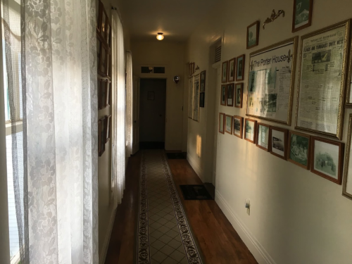 A narrow hallway with white walls, framed photos, and lace curtains, leading to a door at the end.