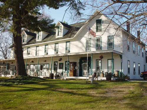A historic white building with green trim, featuring a porch and large windows, surrounded by grass and trees.