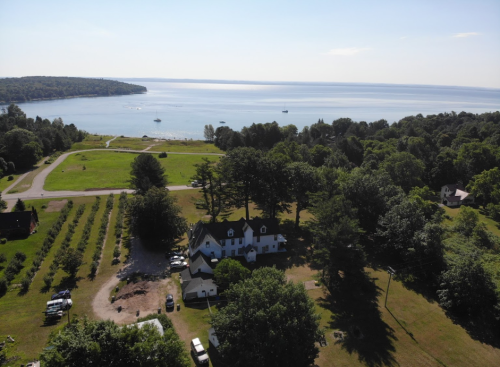 Aerial view of a house near a lake, surrounded by trees and fields, with boats visible on the water.