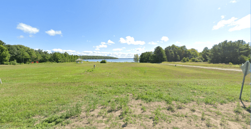 A grassy field with trees in the background, leading to a lake under a blue sky with scattered clouds.