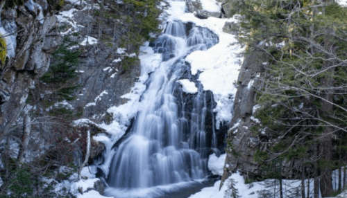 A serene waterfall cascading over rocks, surrounded by snow and evergreen trees in a winter landscape.
