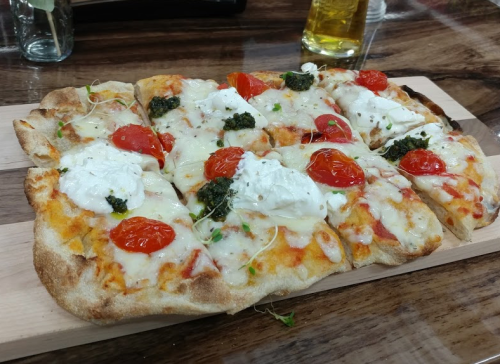 A freshly baked pizza topped with melted cheese, cherry tomatoes, pesto, and herbs, served on a wooden board.