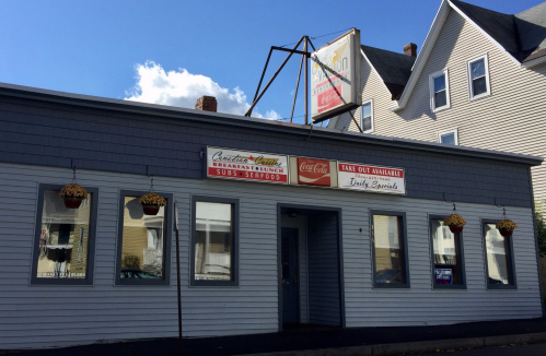 A gray building with signs for a restaurant offering breakfast, subs, and takeout, featuring hanging flower pots.