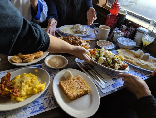 A table filled with various breakfast dishes, including eggs, bacon, pancakes, and a salad, with people reaching for food.