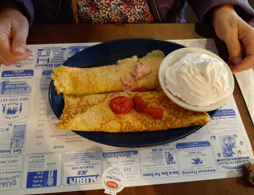 A plate with two crepes filled with fruit, topped with whipped cream, on a printed blue placemat. Hands are visible.