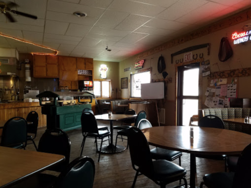 Interior of a casual dining space with wooden tables, chairs, and a bar area, featuring neon signs and a cozy atmosphere.