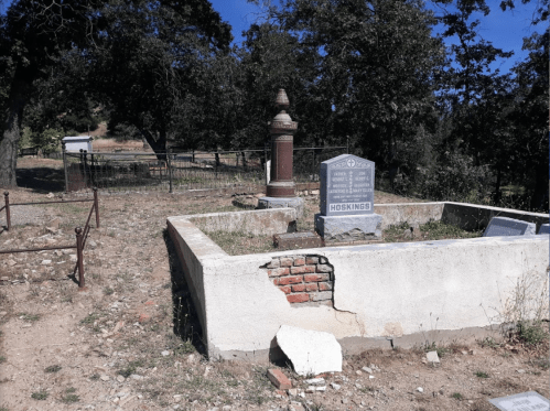 A weathered grave site with a stone marker and a decorative pillar, surrounded by sparse vegetation and trees.
