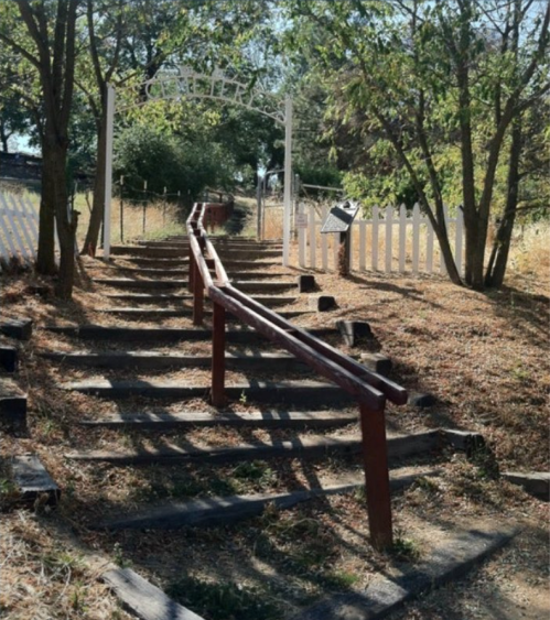 A winding wooden staircase leads up through a grassy area, framed by trees and a white picket fence.