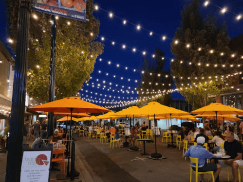 A lively outdoor dining area at night, with yellow umbrellas, string lights, and people enjoying their meals.