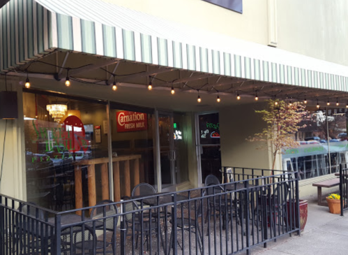 Exterior of a café with a striped awning, string lights, and outdoor seating along a railing.