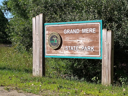 Sign for Grand Mere State Park, featuring a wooden design and a circular emblem, surrounded by greenery.