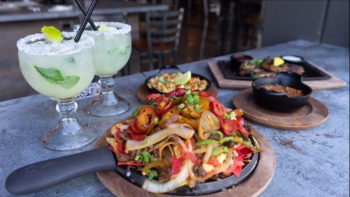 A sizzling platter of fajitas with colorful vegetables, accompanied by two margaritas and a side of rice and beans.