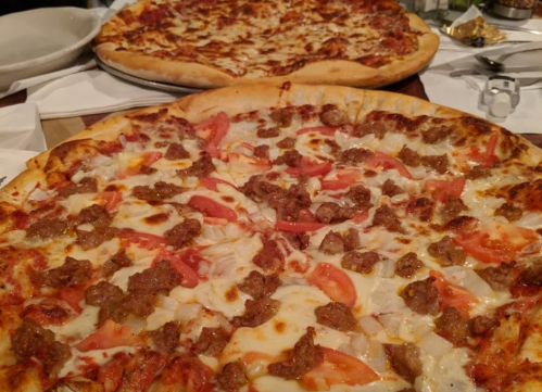 Two pizzas on a table: one topped with sausage, tomatoes, and onions; the other with cheese and a meaty topping.