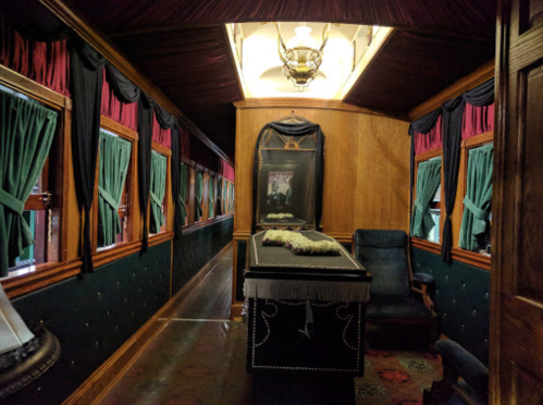 Interior of a vintage train car featuring dark wood paneling, green curtains, and a coffin adorned with flowers.