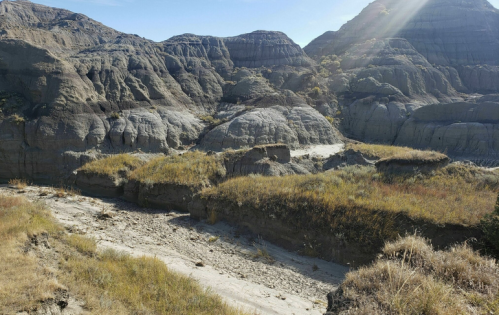 A rugged landscape of gray hills and dry riverbed, with patches of grass under a clear blue sky.